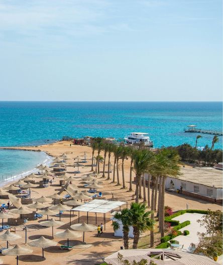 Sandstrand eines Urlaubsresorts am Roten Meer mit Palmen, Reet-Sonnenschirmen und Liegen, im Hintergrund türkisfarbenes Wasser und ein Boot.