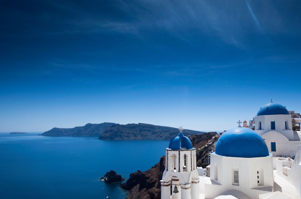 Weiße Gebäude mit blauen Kuppeln in Oia auf Santorin, gelegen an einer Steilküste über dem tiefblauen Ägäischen Meer.