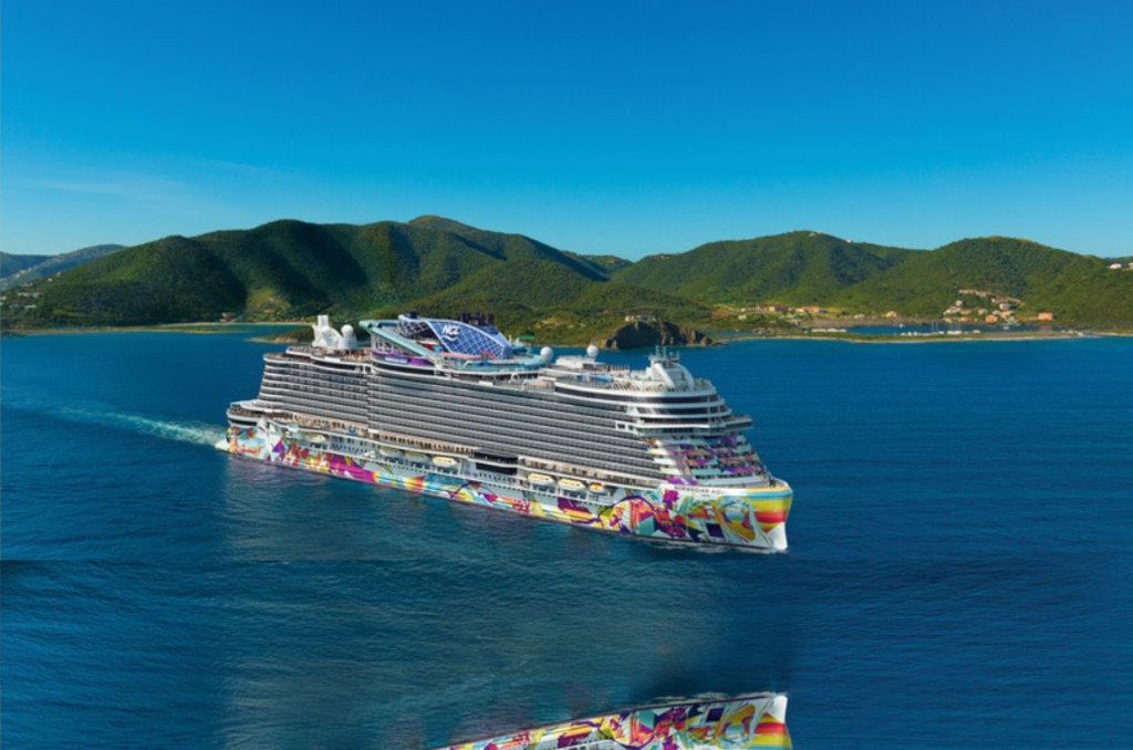 Das Kreuzfahrtschiff Norwegian Aqua mit bunt bemaltem Rumpf fährt auf blauem Meer vor einer grünen Inselkulisse.